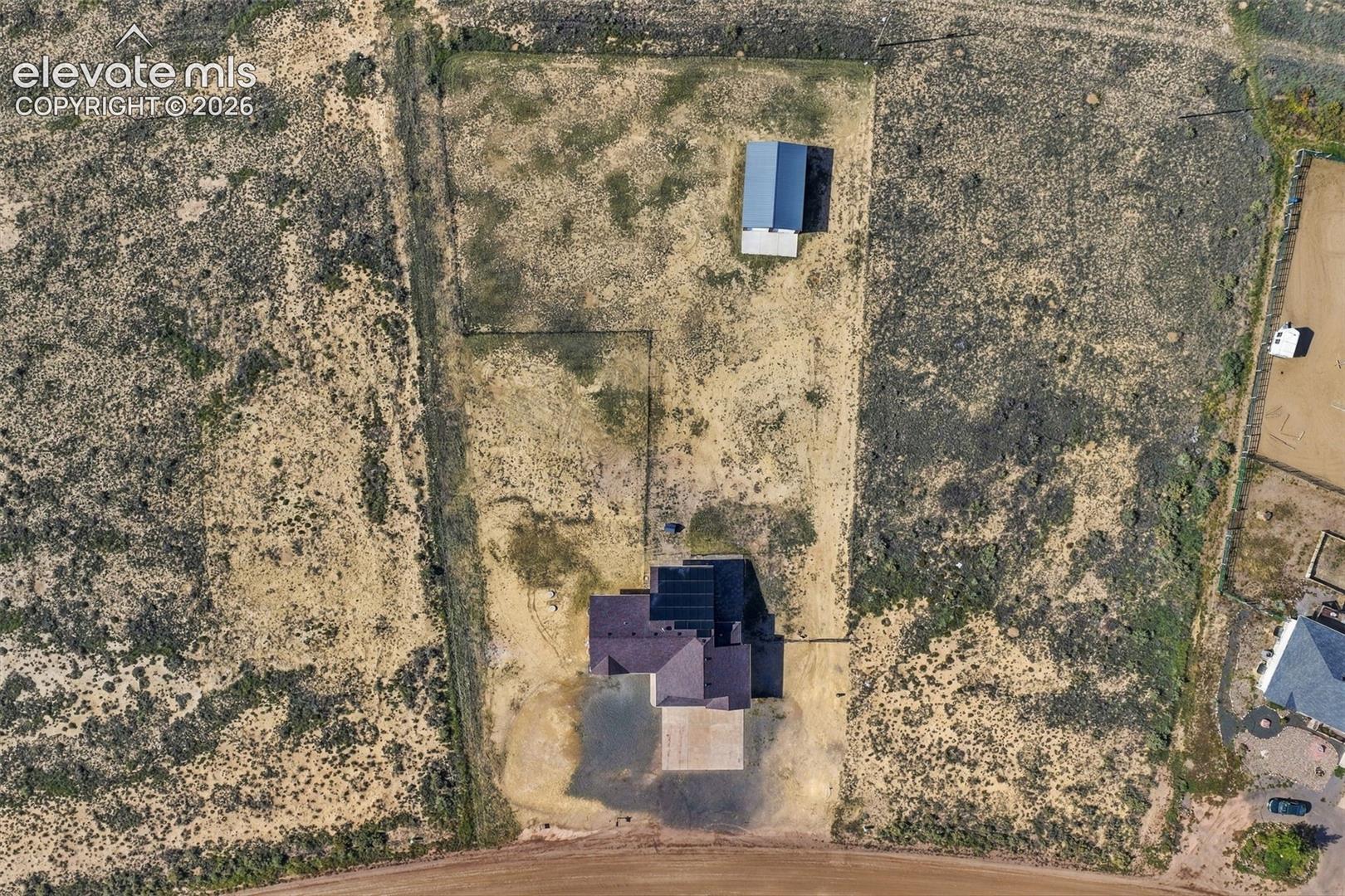 Aerial perspective of a sprawling lot featuring a detached structure with a metal roof, an expansive open land area, and perimeter fencing