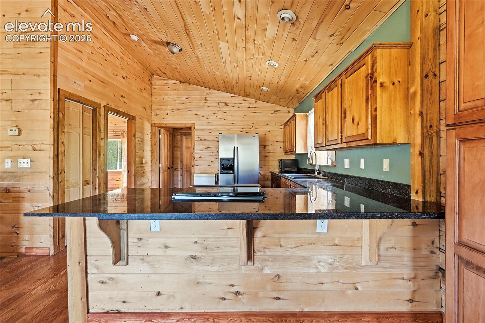 Vaulted wood-paneled ceiling, wood-finish walls, wood-finish flooring, dark stone countertops, and stainless steel refrigerator