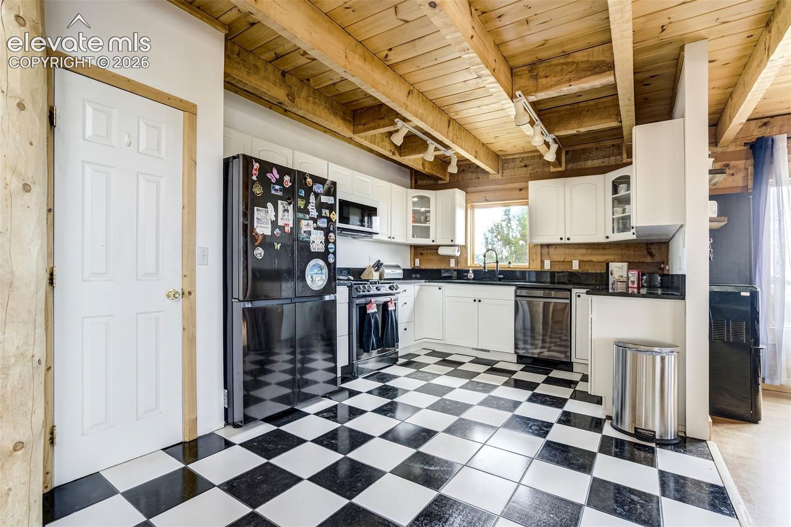 Kitchen featuring exposed wood beam ceilings, white cabinetry, black countertops, a stainless steel dishwasher, and checkerboard floor tiles