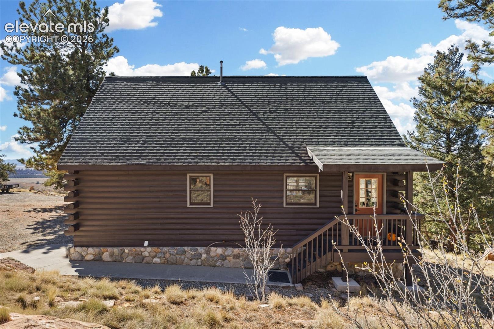 Log construction exterior featuring a stone masonry foundation, shingle roof, covered front porch with railing, and a framed entry door