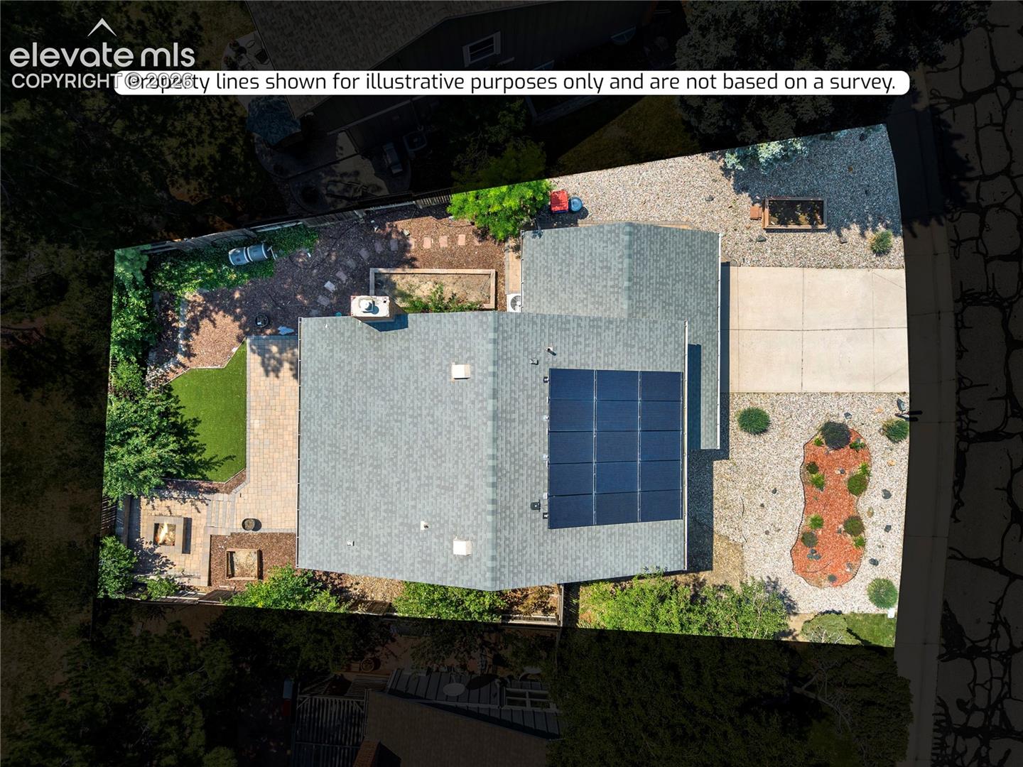 Aerial perspective of a property featuring roof-mounted solar panels, a paved driveway, a hardscaped rear patio area, and a stone-lined fire pit