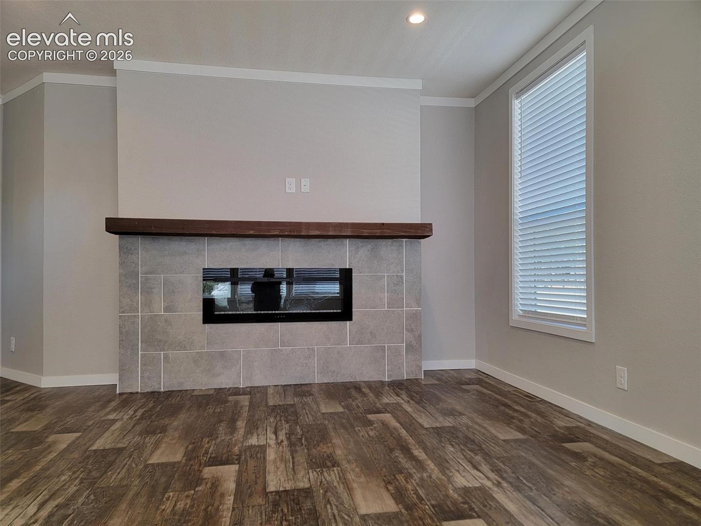 Fireplace featuring a large-format tile surround, recessed electric insert, and a solid wood mantel, complemented by wood-finish flooring and a single tall window with integrated blinds