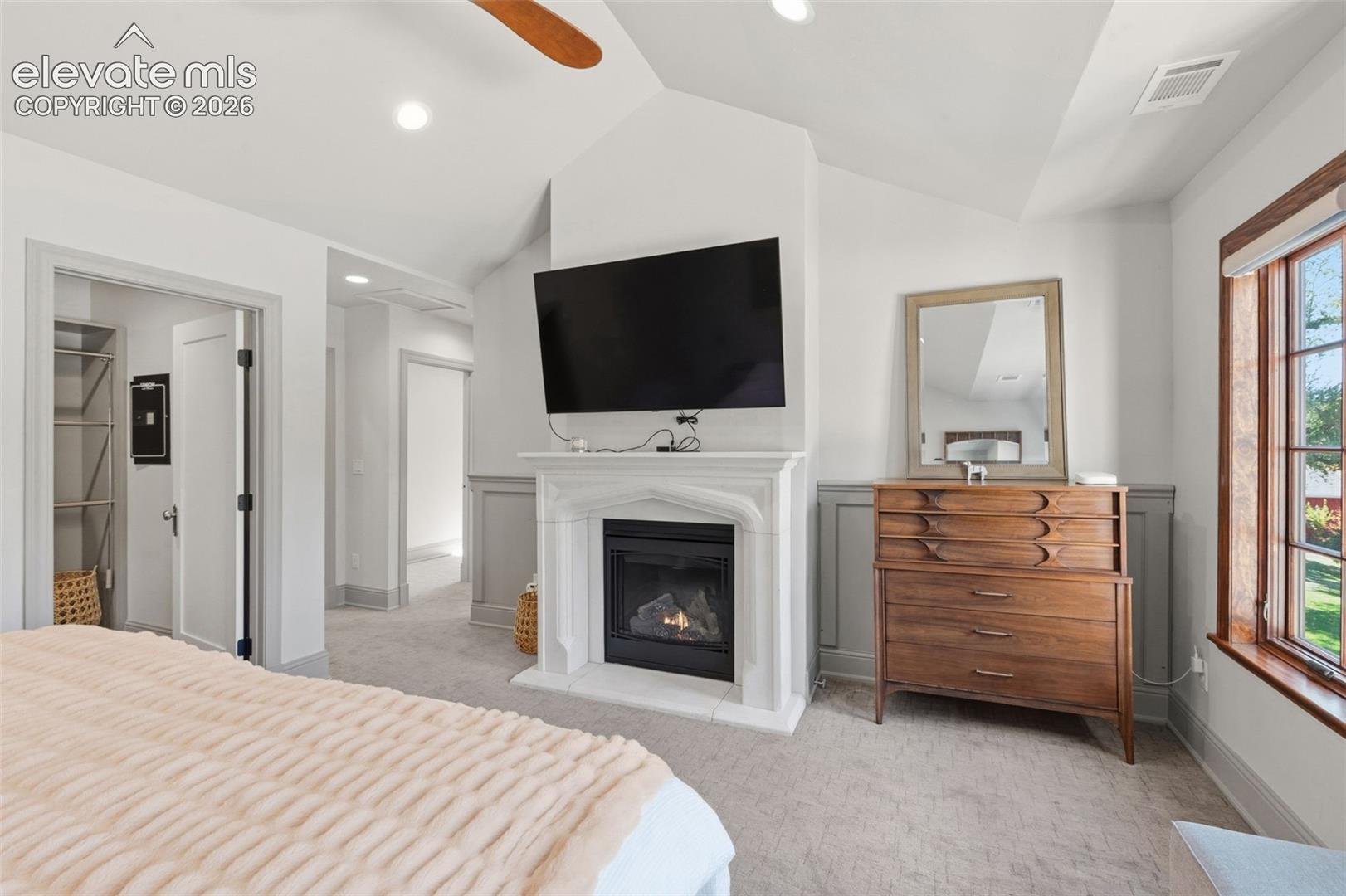 Vaulted ceiling with ceiling fan, fireplace featuring a carved stone-style mantel, wainscoting detail, and multi-pane window with wood trim