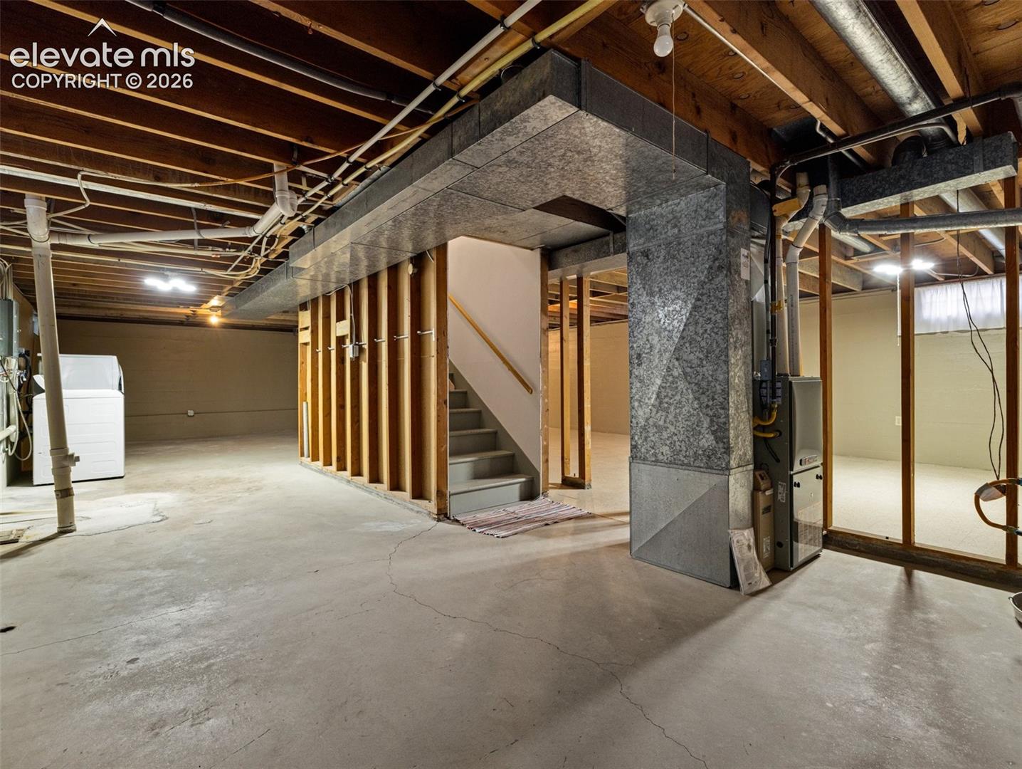 Unfinished basement with concrete flooring and exposed ceiling joists, featuring a utility sink and washer/dryer hookups