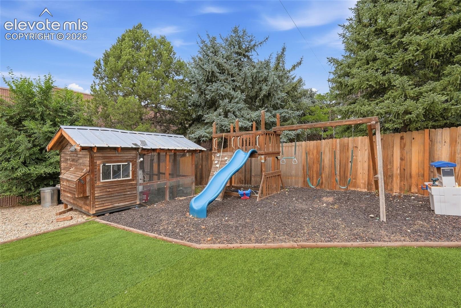 Backyard featuring a wooden privacy fence, well-maintained artificial turf, wood-mulch ground cover, a wooden play structure with a slide, and a wooden outbuilding with a metal roof