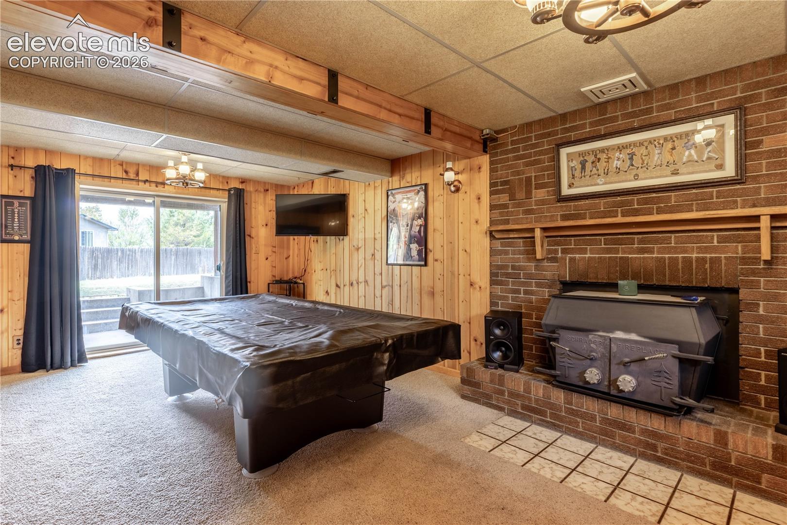Wood-paneled walls featuring exposed ceiling beams, a floor-to-ceiling brick fireplace with a wood-burning insert, and sliding glass doors with exterior access
