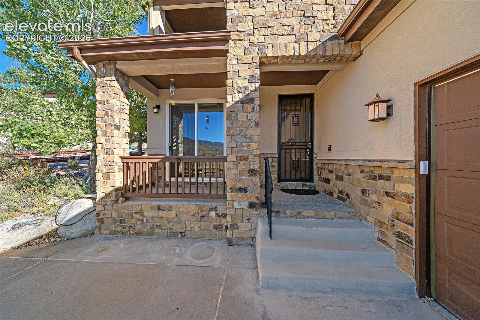 Exterior facade featuring stone veneer siding, a covered entryway with concrete stairs, a metal security screen door, wood-finish railing, and wall-mounted light fixtures
