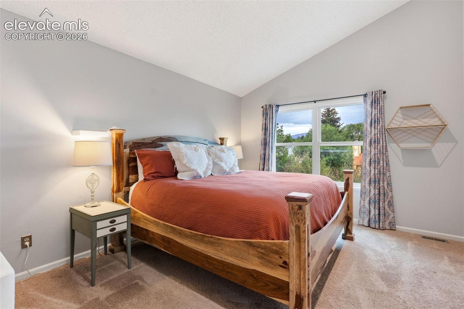 Vaulted ceiling, large window, natural wood bed frame, light gray walls, and carpet flooring