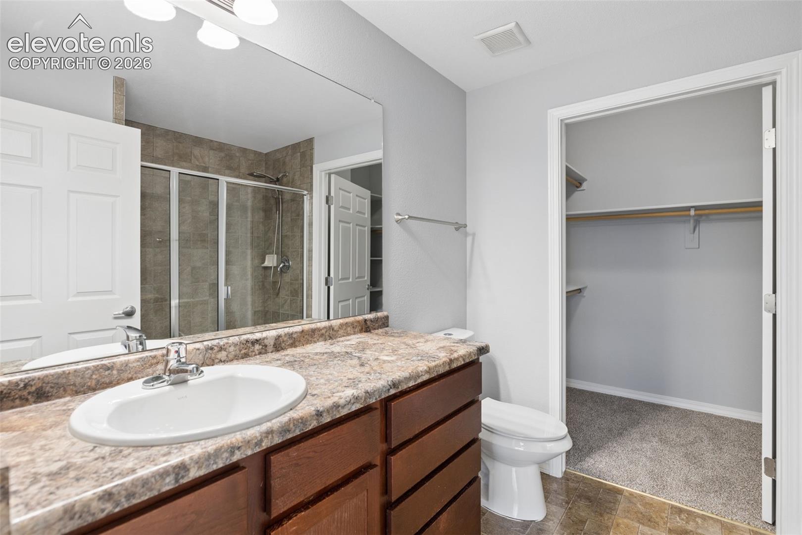 Vanity featuring a stone-patterned countertop, integrated basin, and dark wood cabinetry, paired with a glass-enclosed shower with stone-tile surround and a walk-in closet with built-in shelving