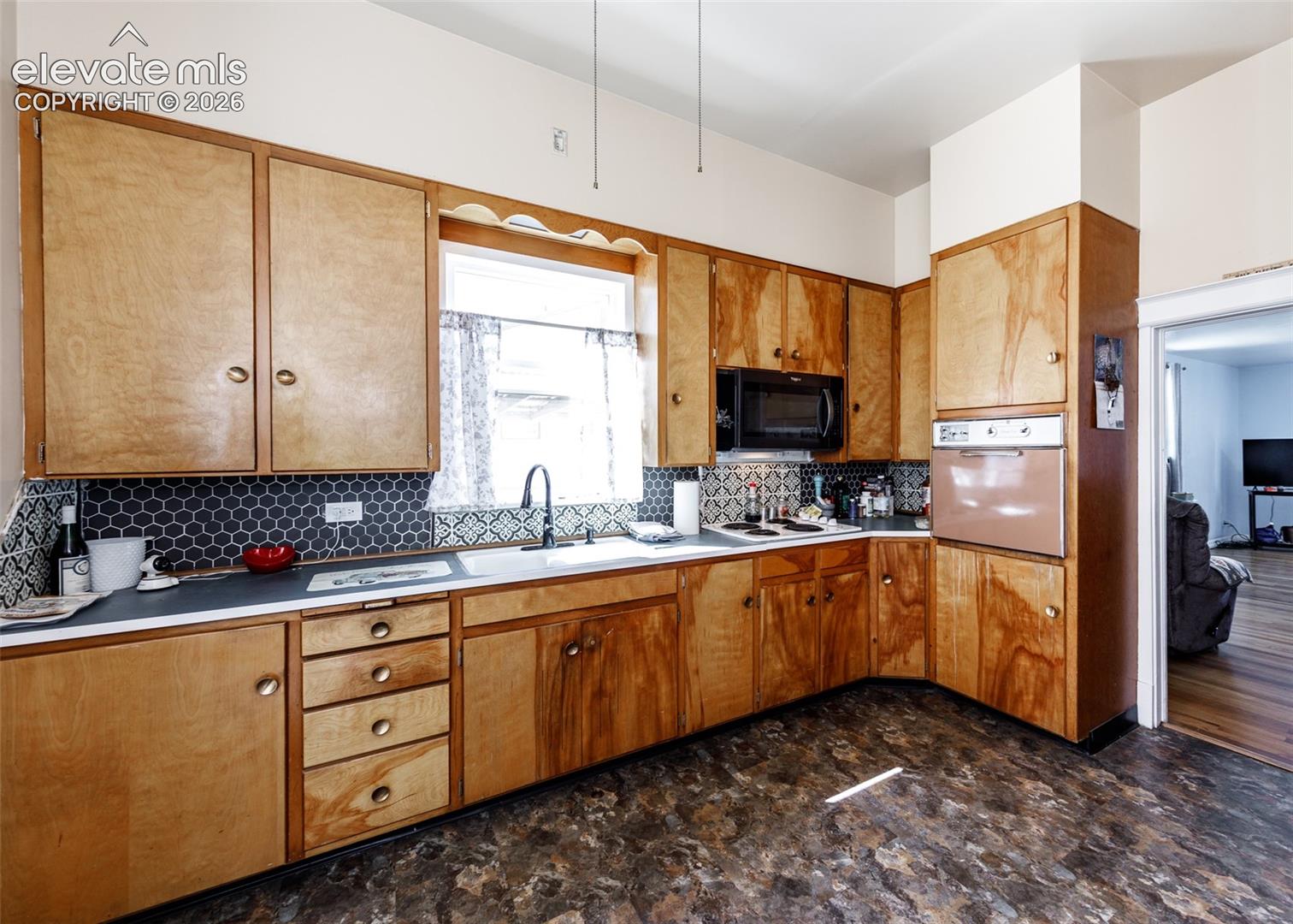 Wood-finish cabinetry with brass-tone hardware, dark hexagonal tile backsplash, white undermount sink with a matte black faucet, built-in microwave, and wood-finish flooring