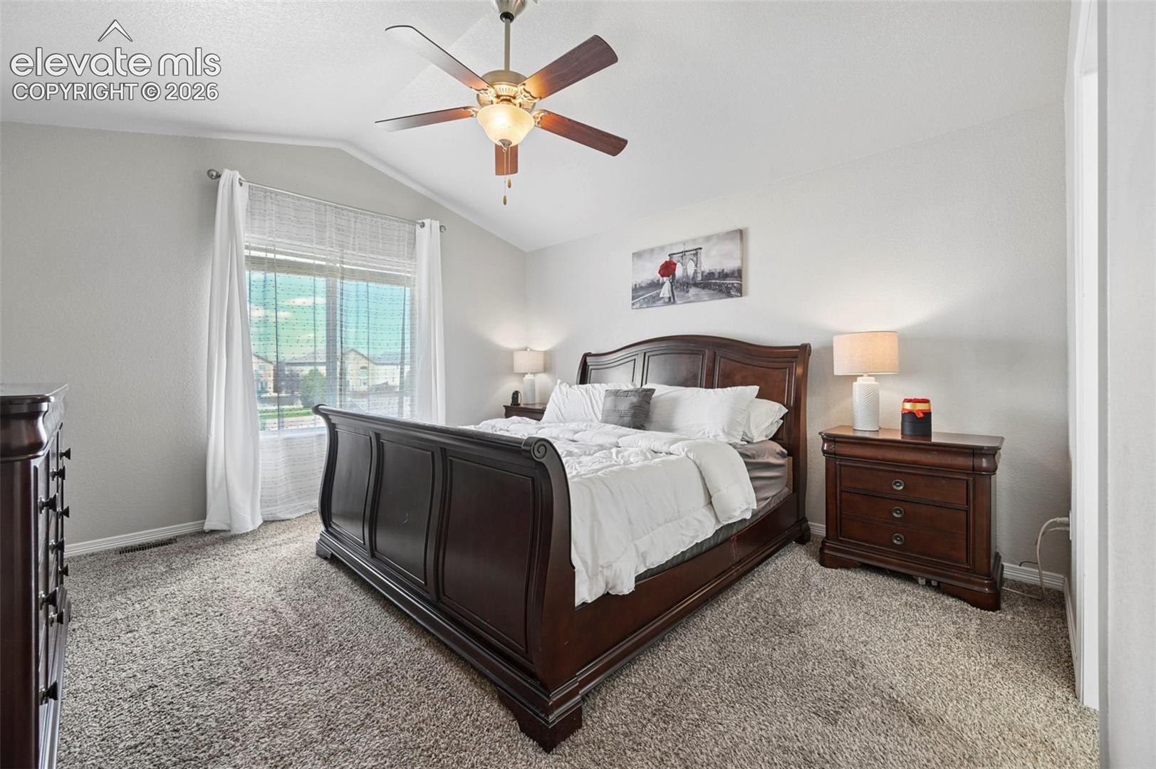 Vaulted ceiling, ceiling fan with light fixture, large window, neutral wall paint, and gray carpet