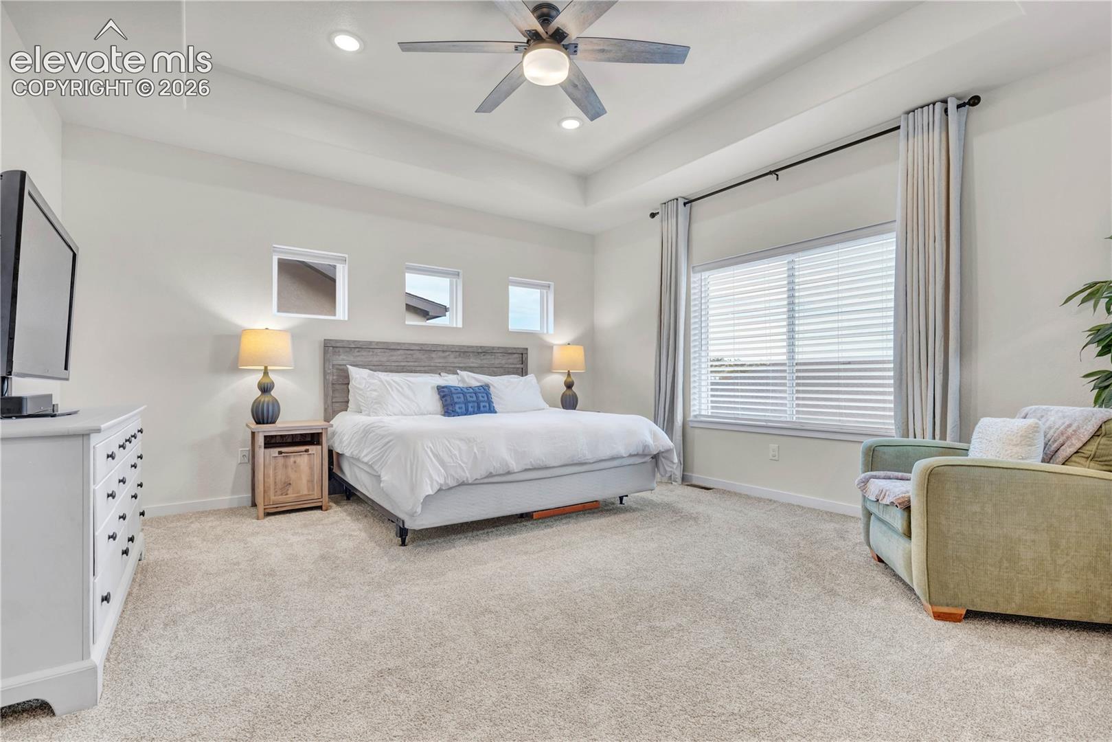 Spacious primary bedroom showcasing neutral carpeting, a ceiling fan with integrated lighting, recessed ceiling fixtures, and large windows with plantation shutters and floor-length drapery