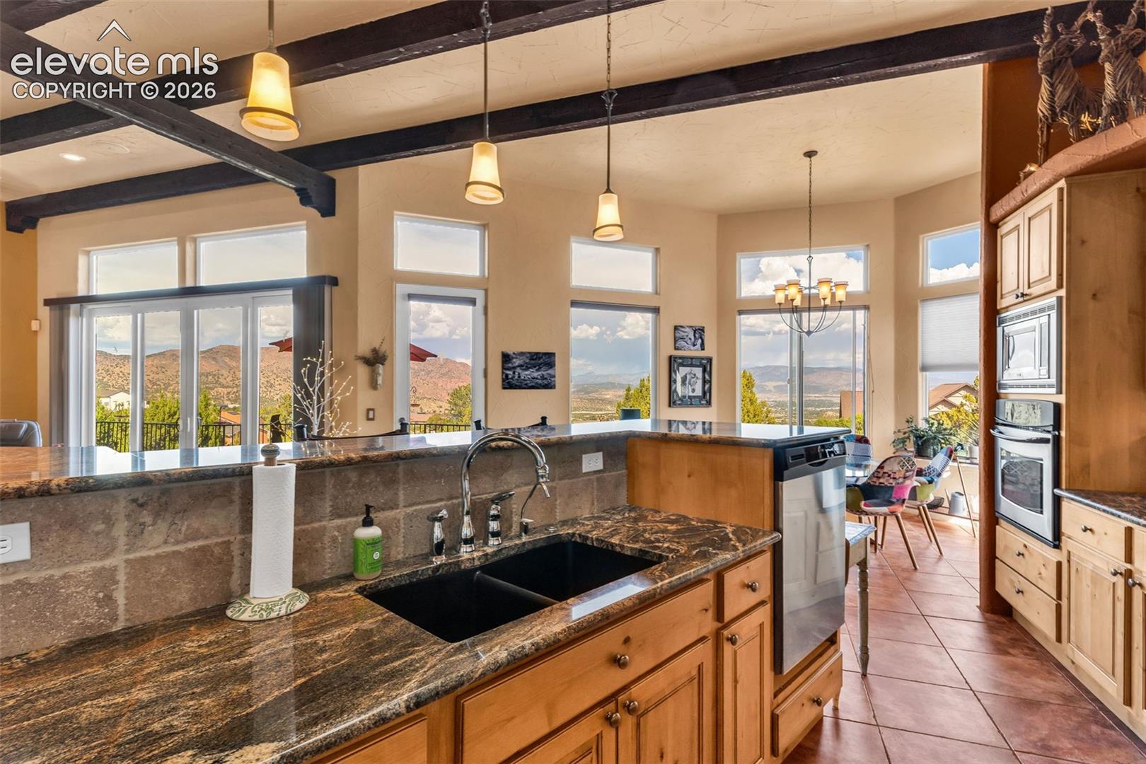 Kitchen featuring natural stone countertops, custom wood cabinetry, dark dual-basin sink with arched faucet, exposed wood ceiling beams, and expansive windows with mountain scenery