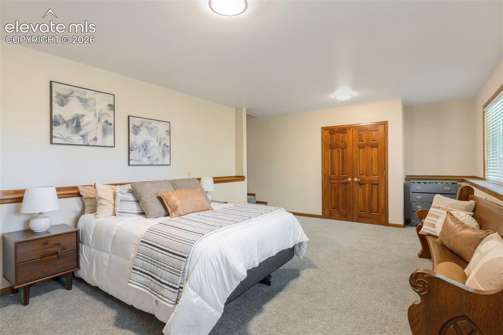 Spacious room featuring light neutral wall tones, plush gray carpeting, built-in wooden bi-fold doors, and a window with horizontal blinds
