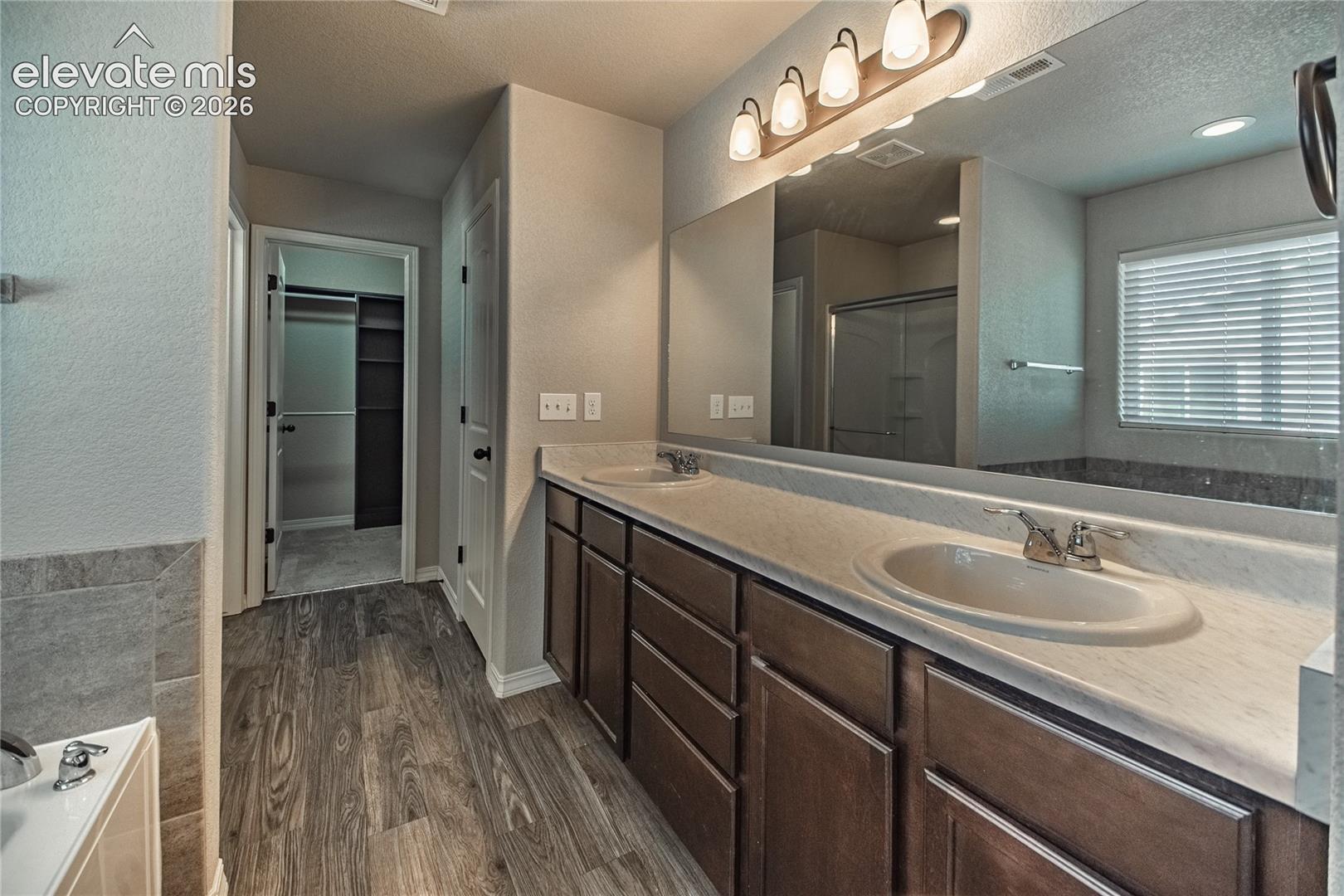 Dual vanity with integrated sinks, wood-finish flooring, a large mirror, and a wall-mounted light fixture