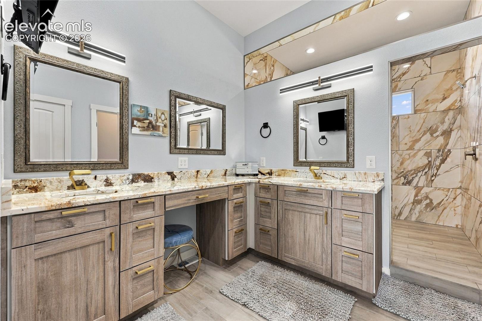 Double vanity featuring wood-finish cabinetry, stone countertops with integrated sinks, brass-toned hardware, framed mirrors, and a walk-in shower with floor-to-ceiling decorative stone-patterned wall tile