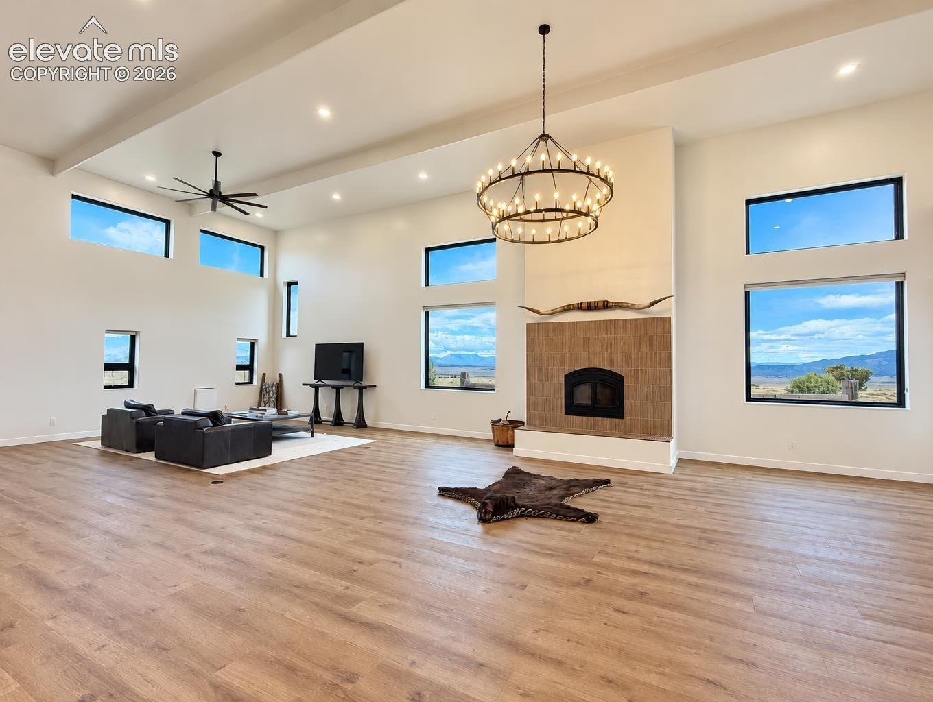 Expansive open-concept room featuring a vaulted ceiling, wood-finish flooring, recessed lighting, a large-scale circular iron chandelier, and a fireplace with a textured tile surround