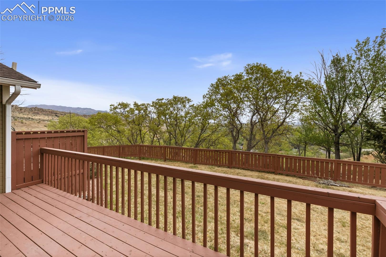 The property features a spacious deck with a brown wooden railing and fencing, overlooking a grassy area with mature trees and distant hills