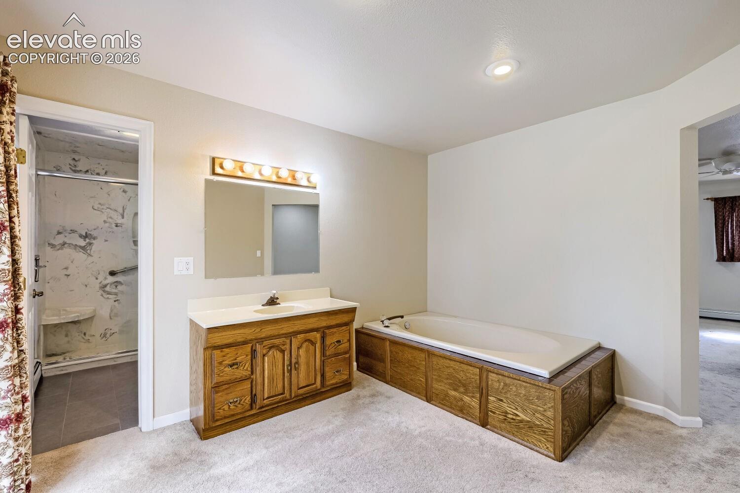 Recessed ceiling light, built-in bathtub, wood-finish vanity with countertop, wall-mounted mirror with overhead lighting, and an adjacent shower enclosure