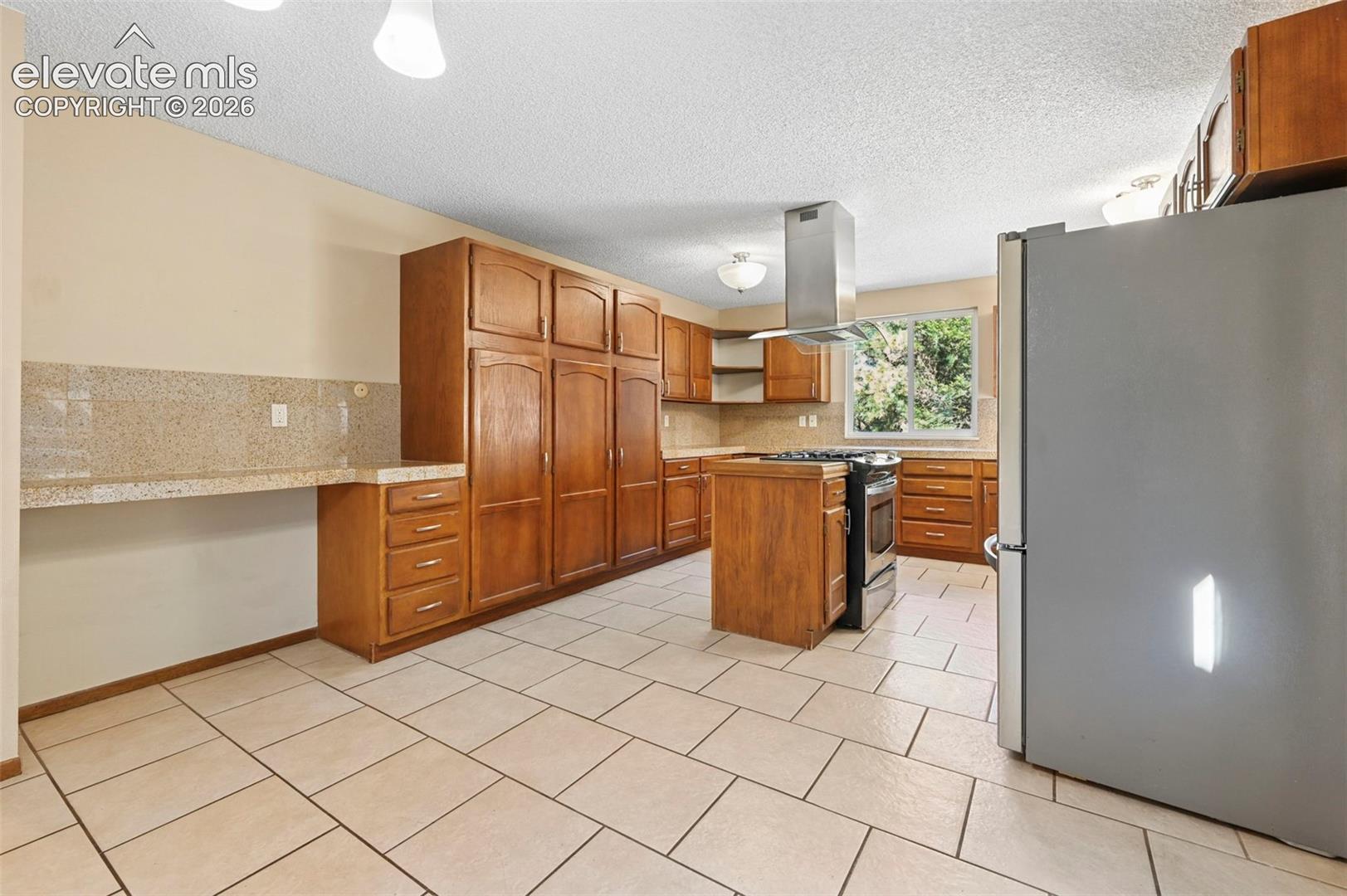 Kitchen featuring natural wood cabinetry, a center island with a gas range, a stainless steel chimney-style range hood, tile flooring, and stone-finish countertops