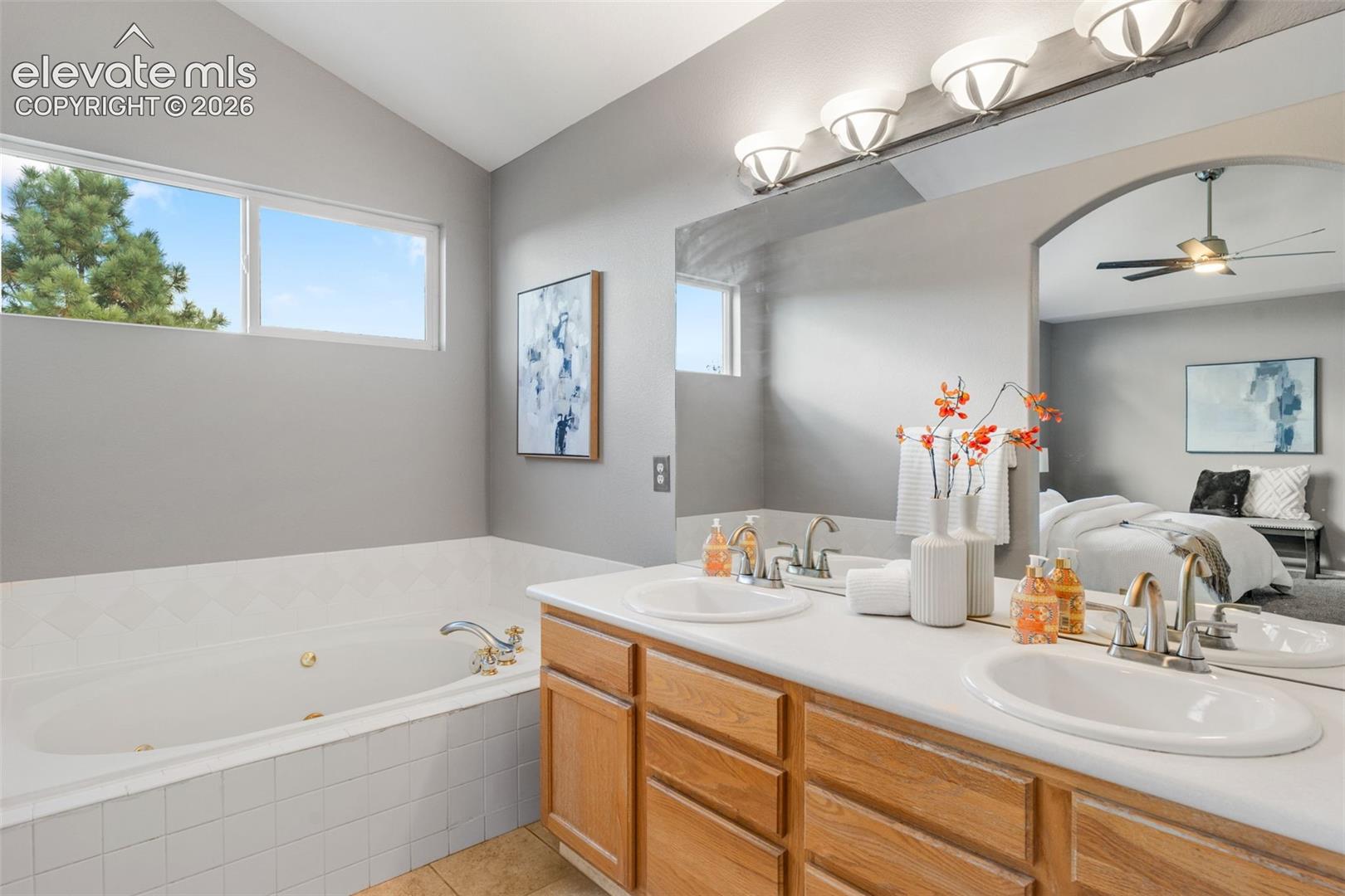 Bathroom featuring a dual-sink vanity with light-tone wood cabinetry, a jetted soaking tub with tile surround, and a large wall-mounted mirror