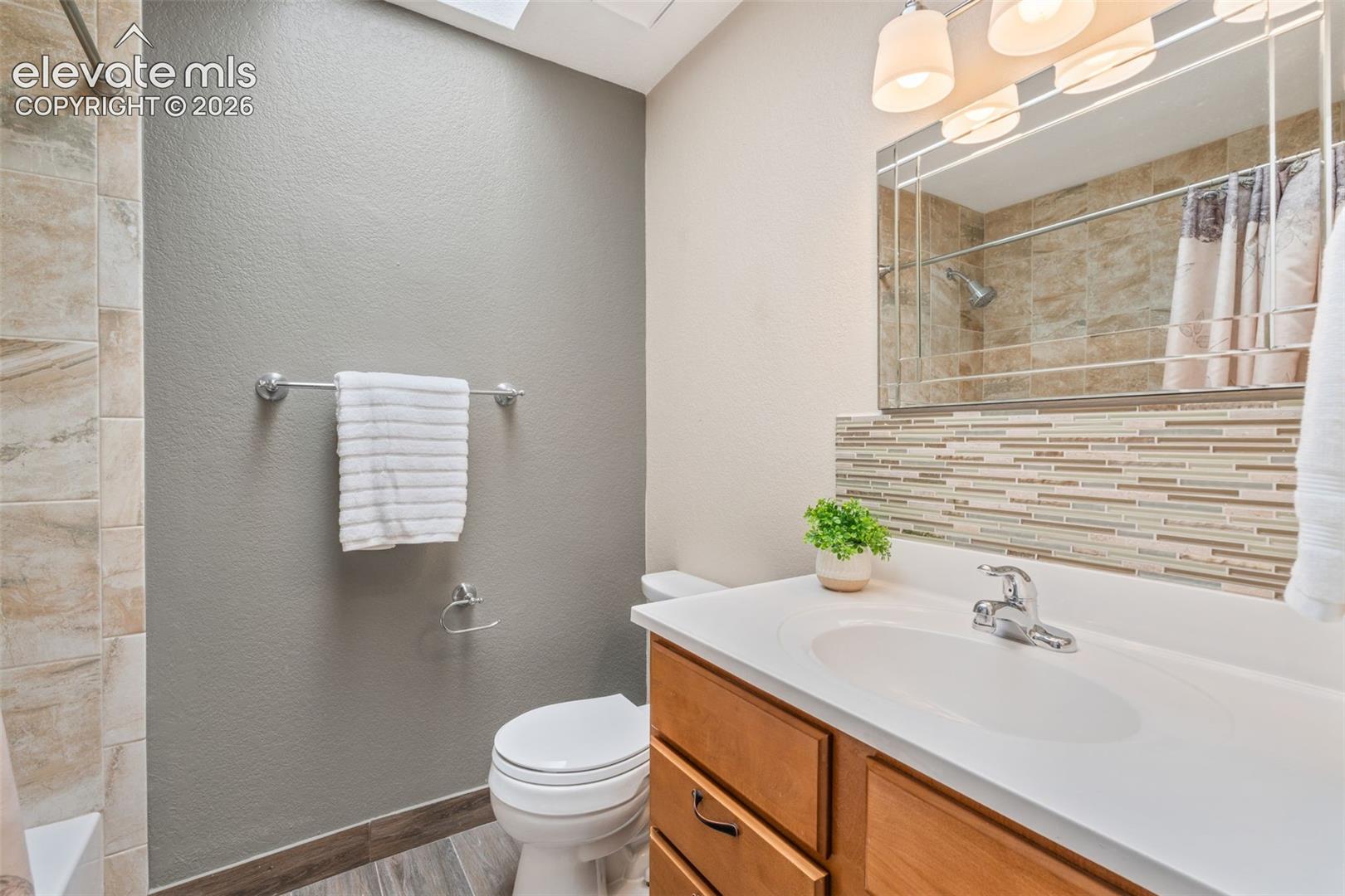 Bathroom featuring a skylight, wood-finish vanity with a white countertop, integrated sink, decorative linear tile backsplash, and a framed vanity mirror with integrated lighting