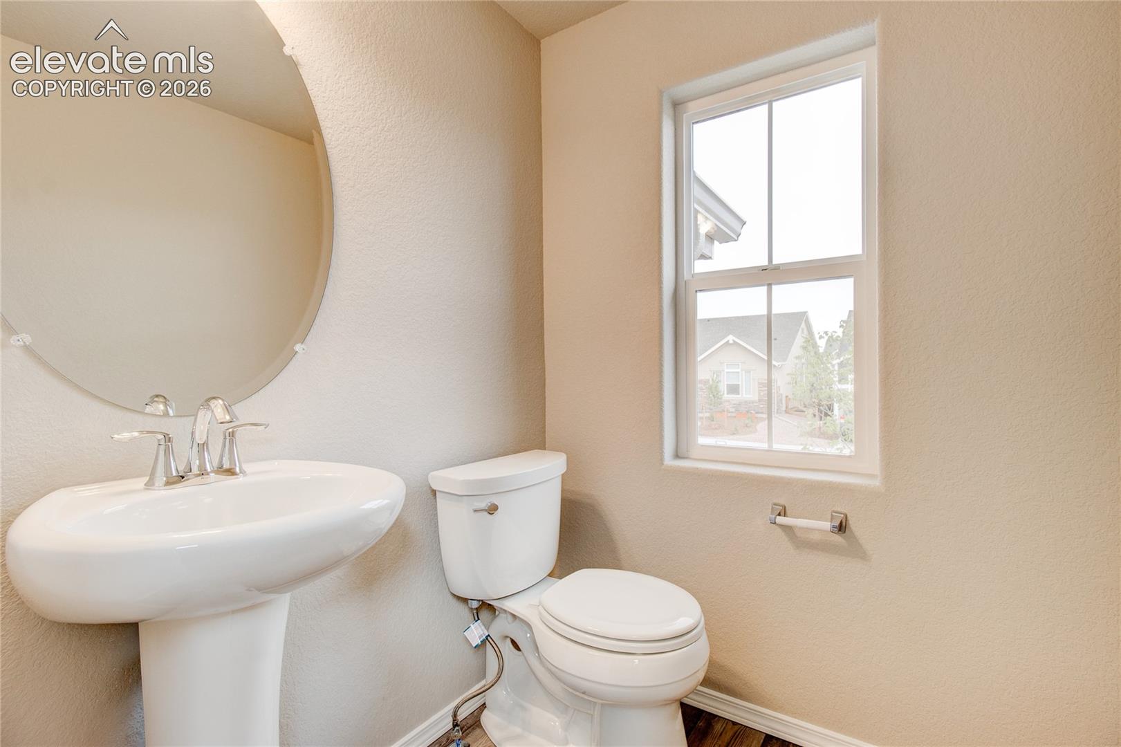 Pedestal sink with a polished chrome faucet, round wall-mounted mirror, two-piece toilet, and single-hung window with a muntin grid