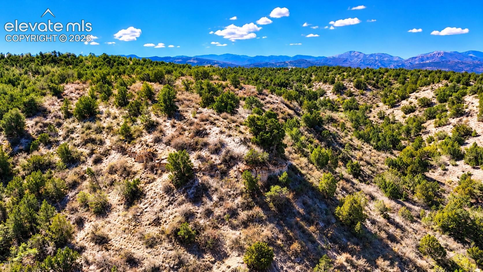 Stunning aerial view of rugged hillsides, native vegetation, and winding country roads, with dramatic mountain peaks extending across the horizon.

