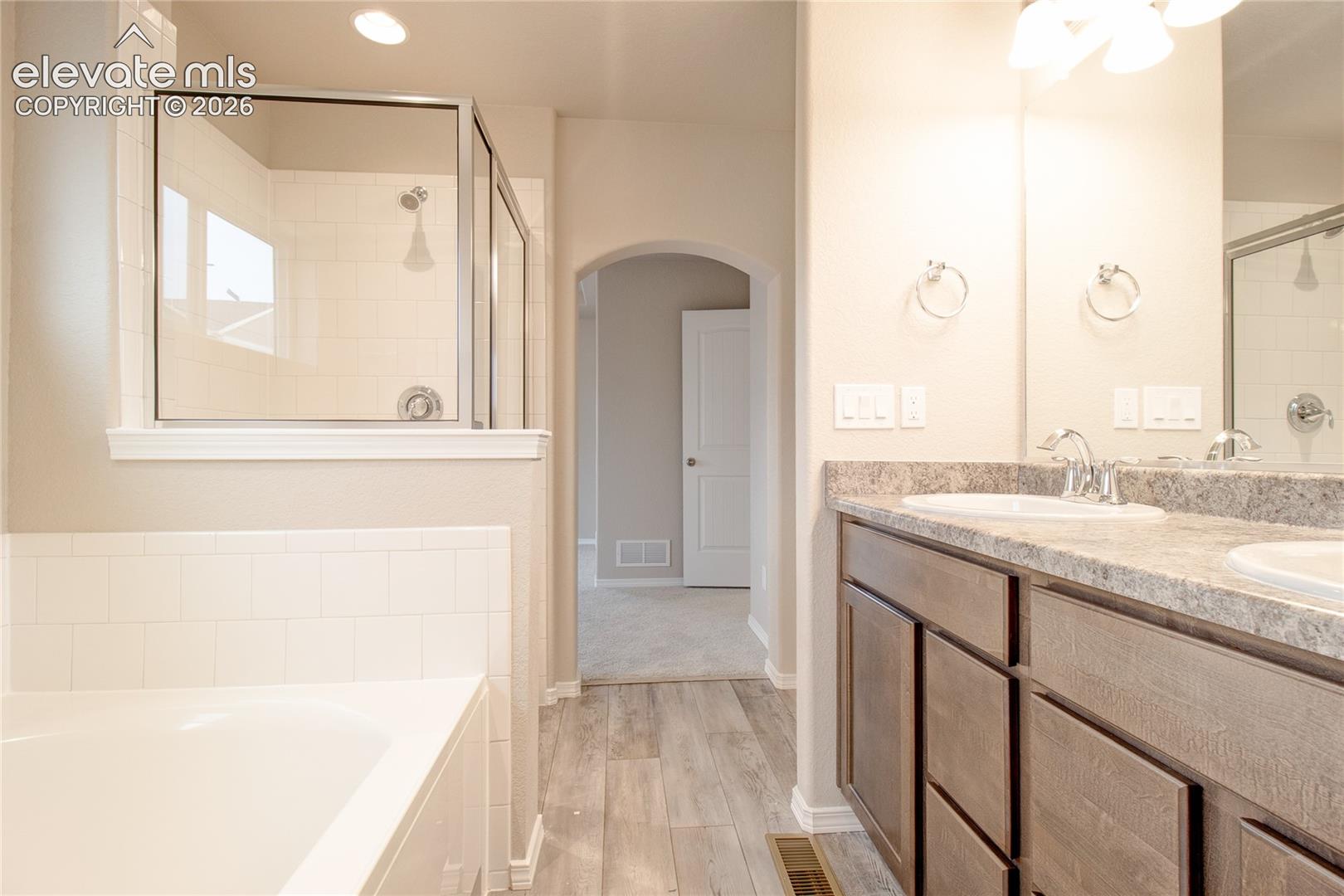 Dual-sink vanity with stone-finish countertop, wood-finish cabinetry, separate soaking tub, and glass-enclosed shower with tiled surround