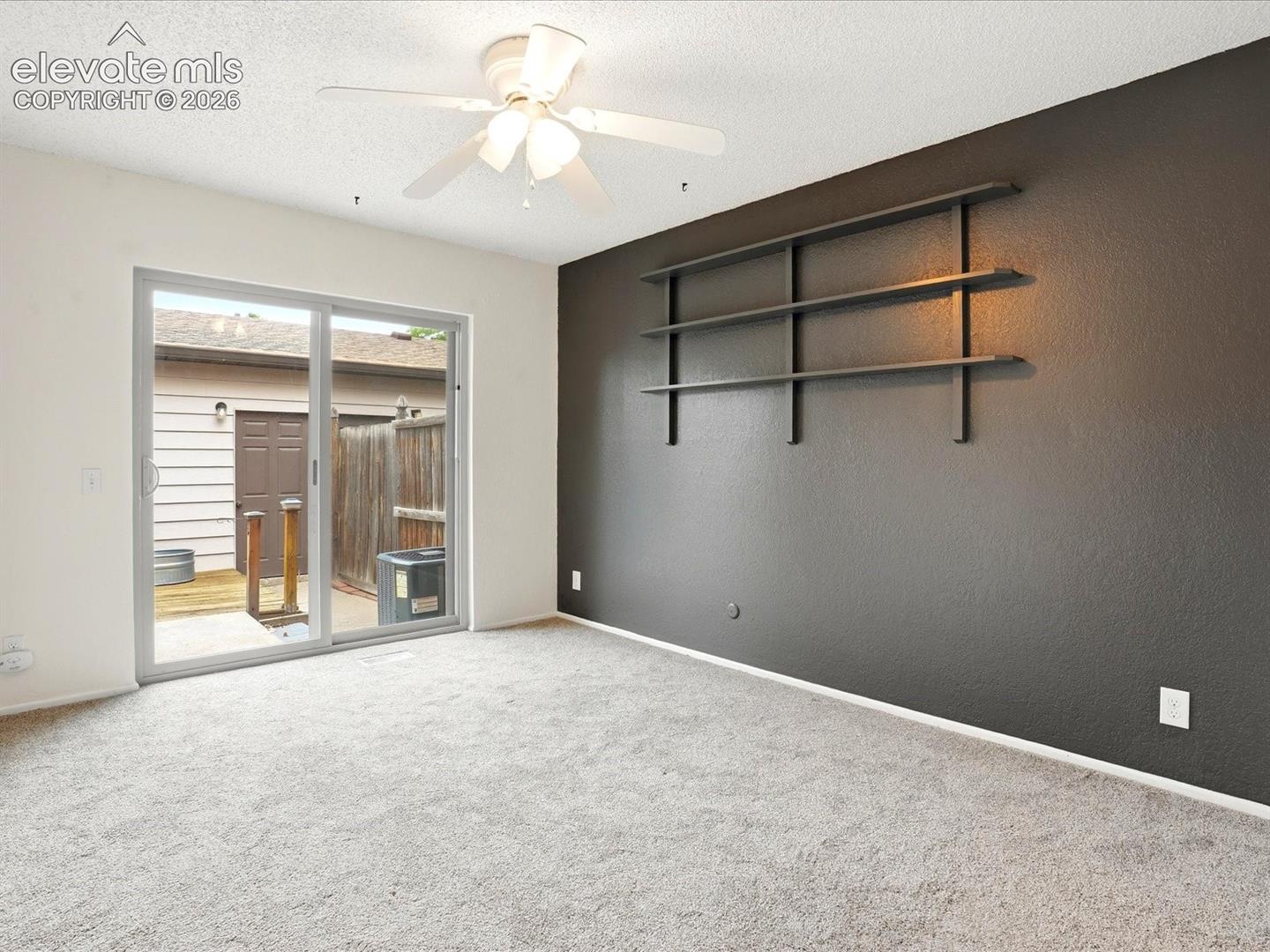 Room featuring carpeted flooring, a dark-toned accent wall, built-in shelving, a ceiling fan with light fixture, and a sliding glass door