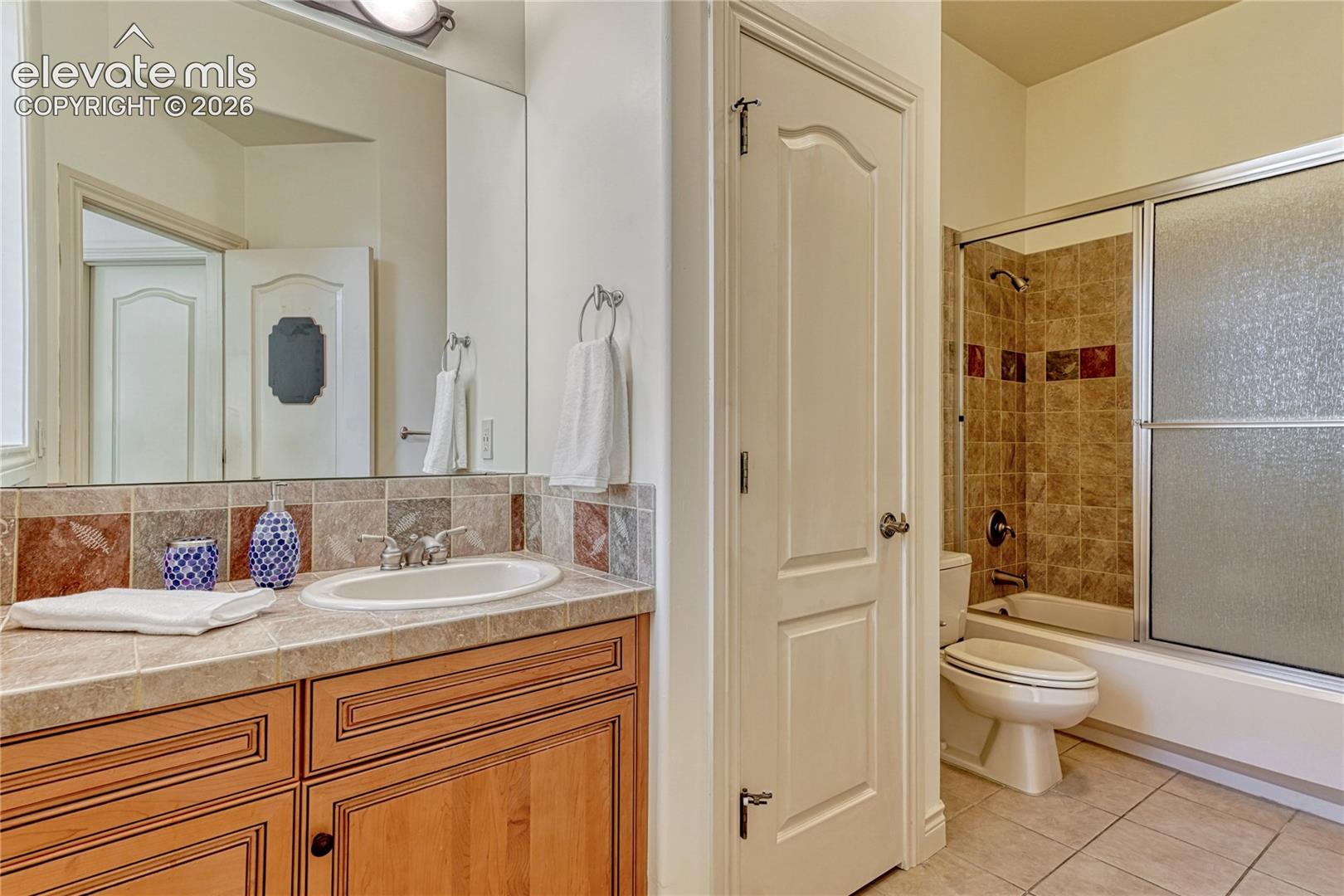 Bathroom featuring a wood-finish vanity with an integrated tile countertop, inset basin, tub-shower combination with sliding glass doors, decorative tile backsplash, and large wall-mounted mirror