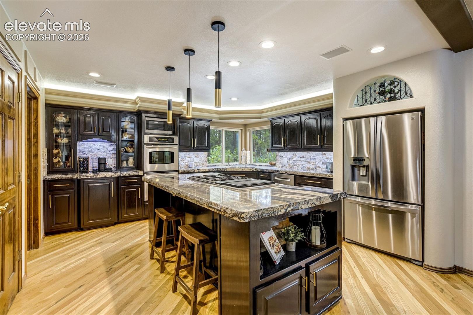Kitchen featuring wood-finish flooring, dark wood cabinetry, stainless steel appliances, a large center island with a cooktop, and granite countertops