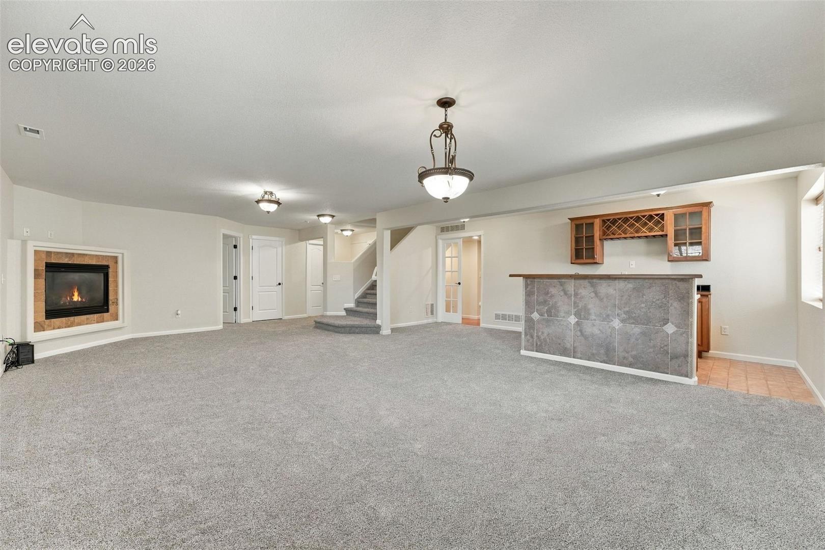 Built-in wet bar featuring stone-tile facing, cabinetry with integrated wine rack, carpeted flooring, fireplace with stone surround, and decorative pendant lighting