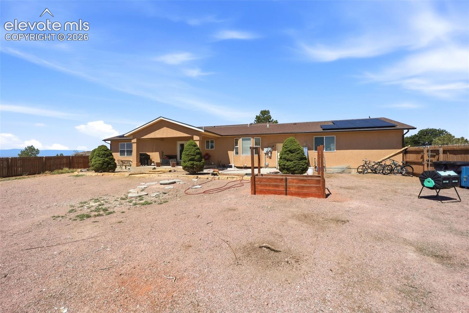 South-facing stucco exterior with a composite shingle roof, integrated solar panels, a wooden privacy fence, and a gravel yard