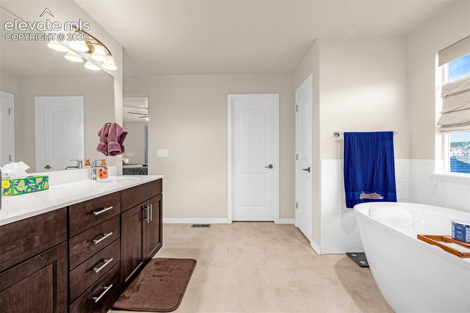 Bathroom featuring dark wood-finish cabinetry, a white countertop with integrated dual sinks, a large wall-mounted mirror, a freestanding soaking tub, and tile flooring