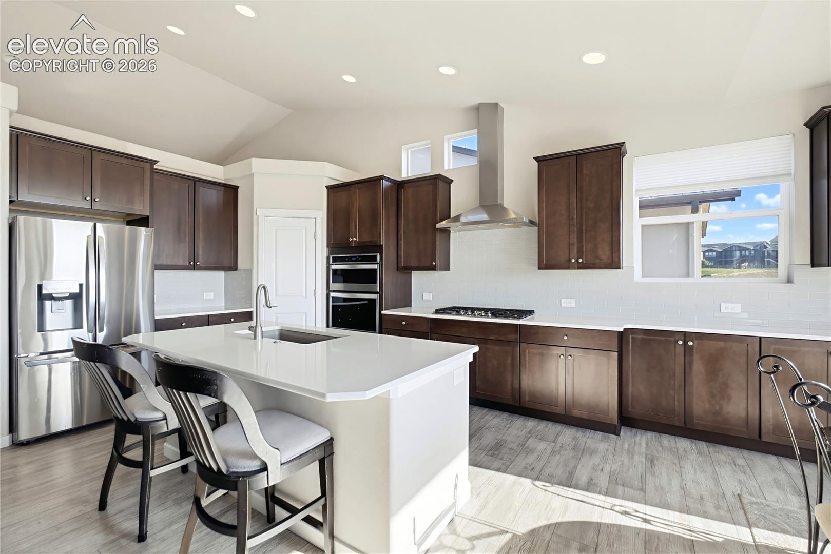 Kitchen featuring dark wood cabinetry, quartz countertops, a central island with an integrated sink, stainless steel appliances, and wood-finish flooring