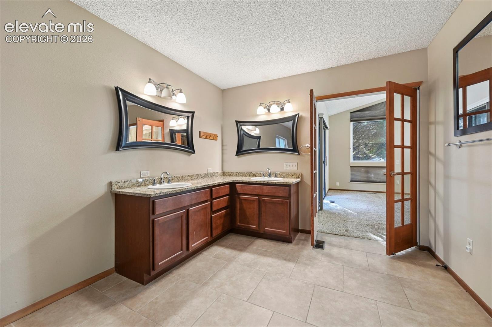 L-shaped vanity with stone-finish countertop, dual under-mount sinks, wood-finish cabinetry, ceramic tile flooring, and French doors