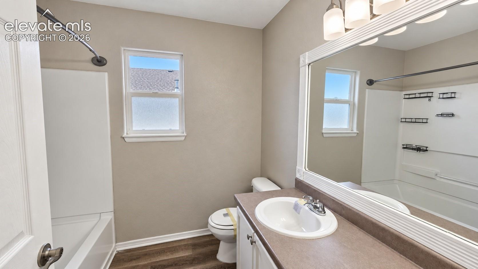 Bathroom featuring wood-finish flooring, a white vanity with a neutral solid-surface countertop, a wide-frame wall mirror, and a tub-shower combination with built-in shelving