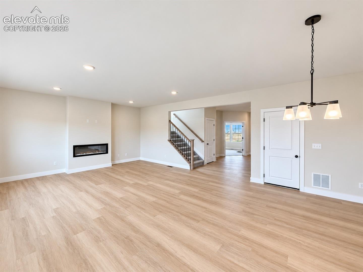 Open-concept interior featuring wood-finish flooring, recessed lighting, a linear wall-mounted fireplace, a tiered chandelier, and a staircase with a wooden handrail