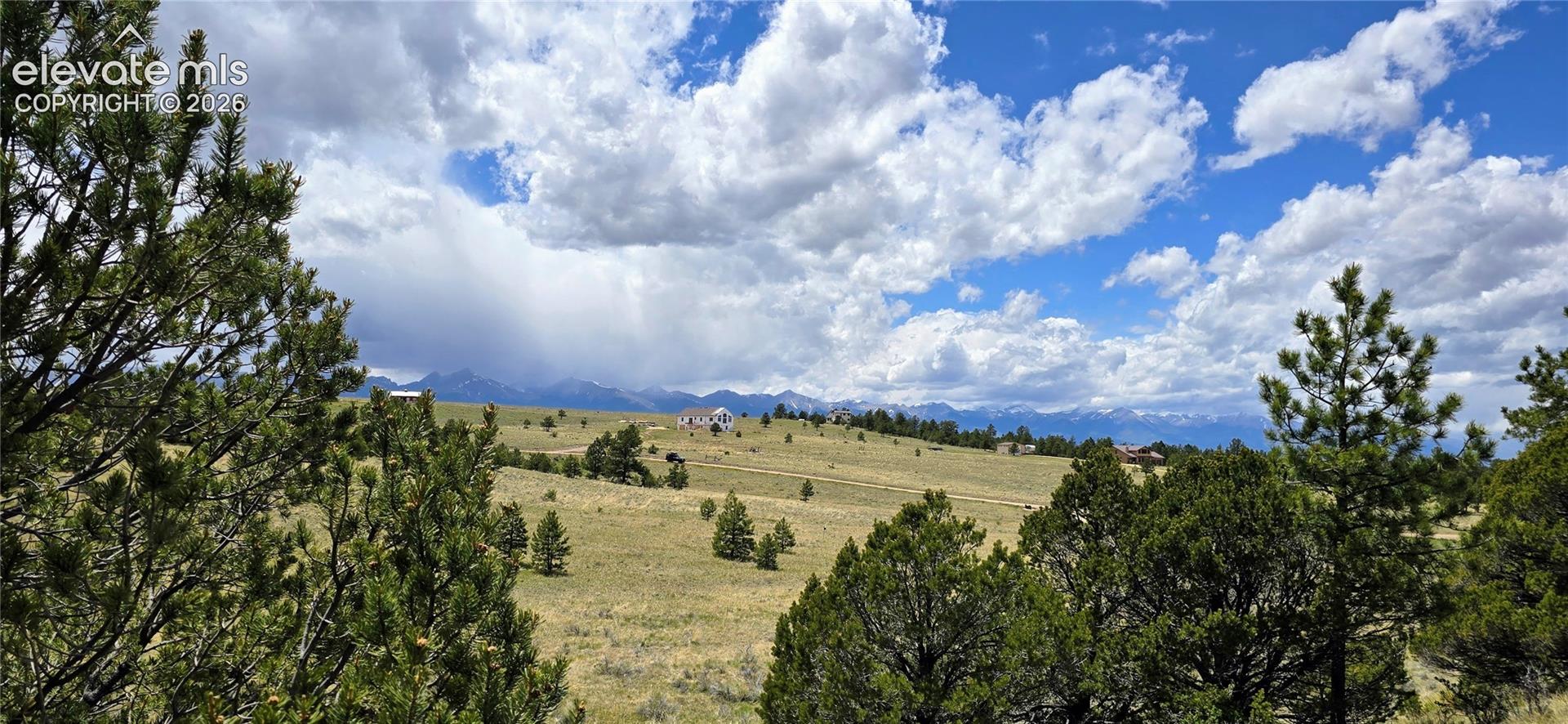 Expansive rolling landscape with native vegetation and scattered evergreen trees, offering panoramic vistas of distant mountain ranges beneath a dramatic cloud-filled sky