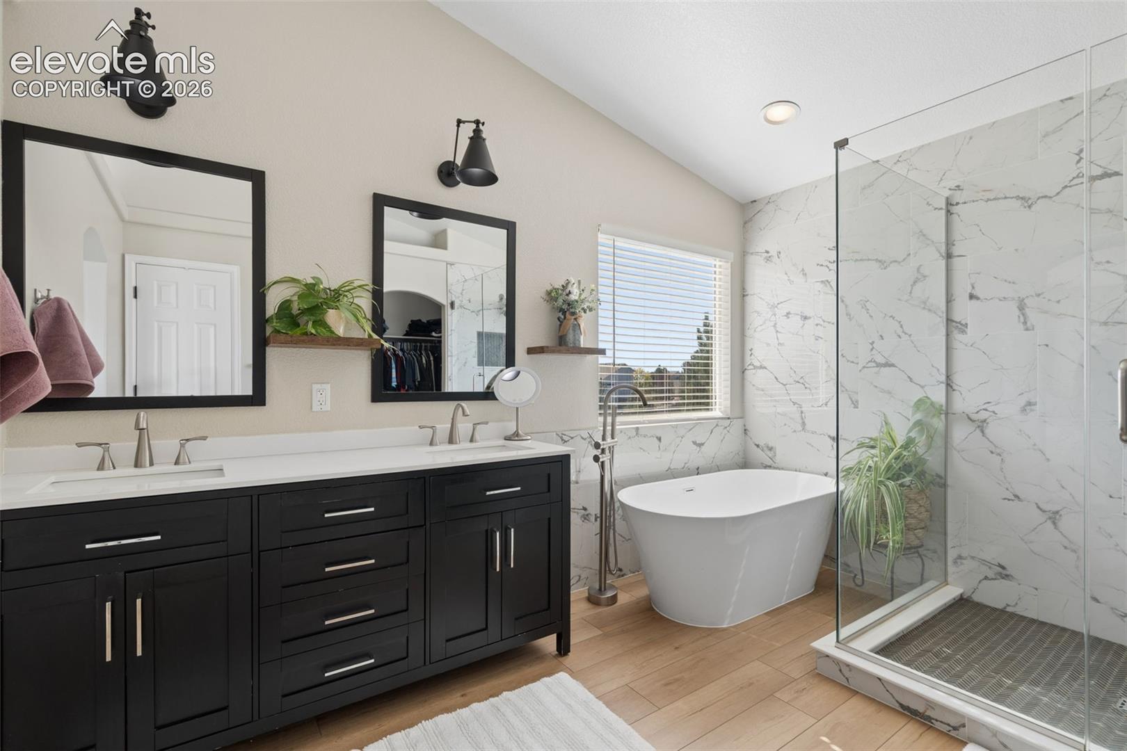 Dual vanity with white countertops, freestanding soaking tub, glass-enclosed shower with tiled walls, luxury vinyl flooring