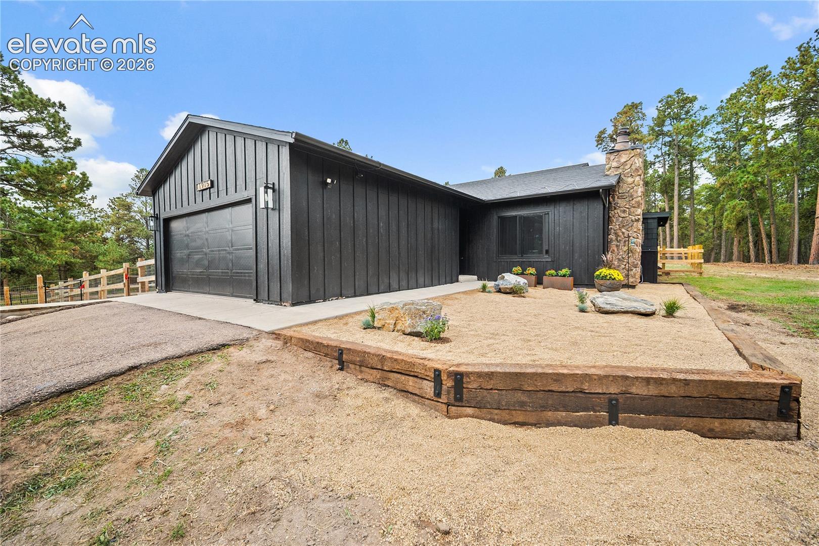 Modern exterior featuring black vertical siding, a stone masonry chimney, a concrete driveway, and a tiered retaining wall constructed of timber railroad ties