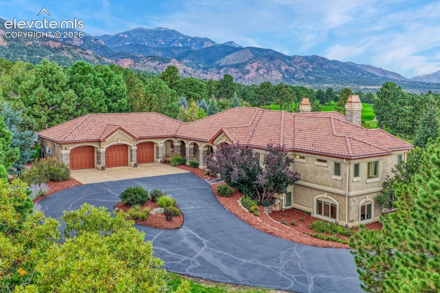 Stucco exterior featuring a multi-gabled clay tile roof, three-car garage, arched entryways, two masonry chimneys, and a mountain backdrop