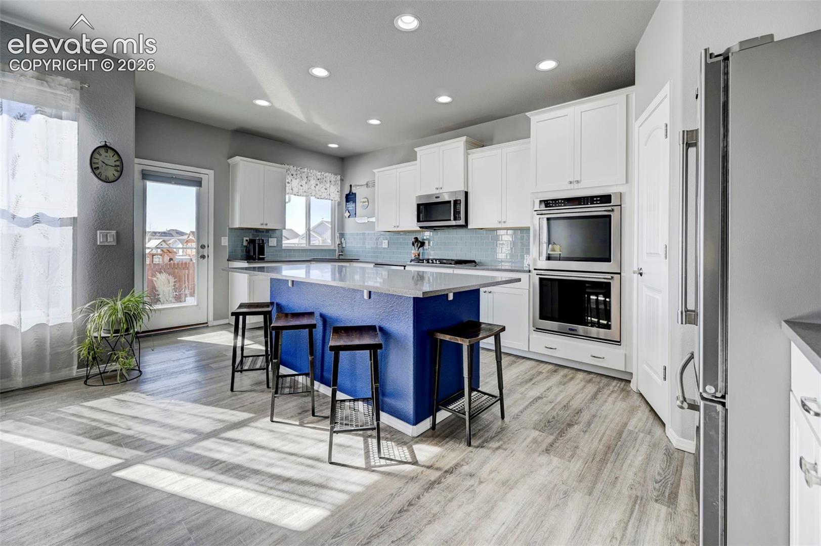 Kitchen featuring a blue island with seating, white cabinetry, built-in stainless steel double ovens, a stainless steel microwave, and wood-finish flooring