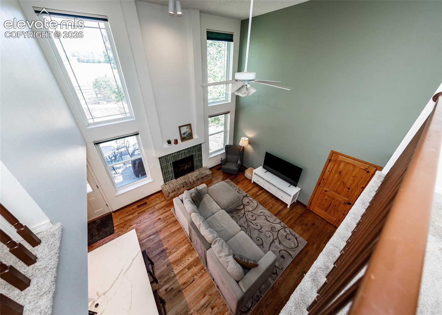 Vaulted ceiling, hardwood flooring, fireplace with brick hearth, multiple tall windows, and a ceiling fan