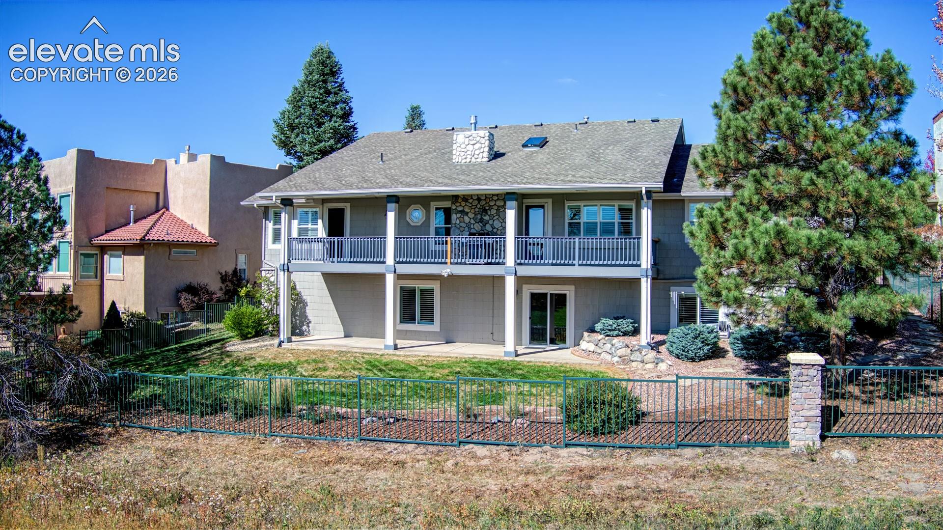 Two-story exterior featuring a stone chimney, a full-width upper-level balcony with railings, a lower-level concrete patio, and fenced-in grounds with mature pine trees
