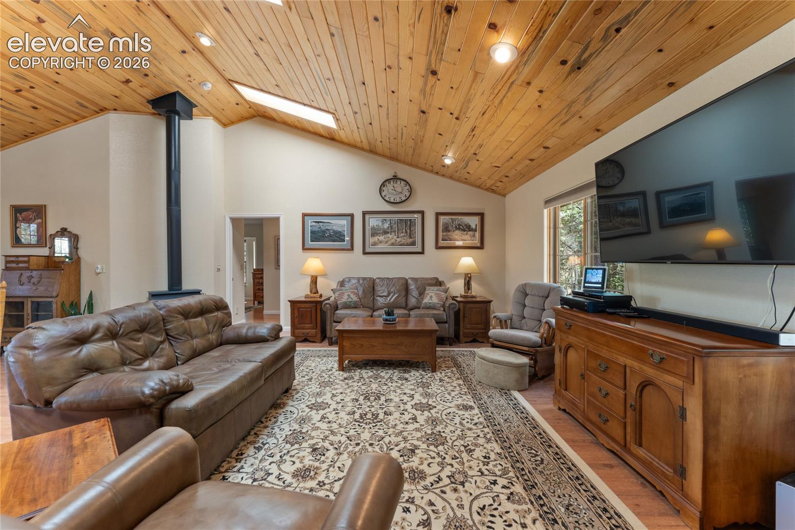 Vaulted wood-paneled ceiling with skylights and recessed lighting, featuring a prominent stand-alone stove with black stovepipe, light-toned wall finishes, and wood-finish flooring