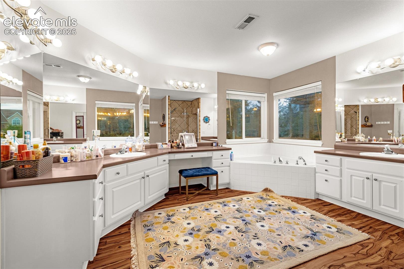 Dual vanity with white cabinetry, built-in makeup desk, large soaking tub, wood-finish flooring, and multiple mirrors