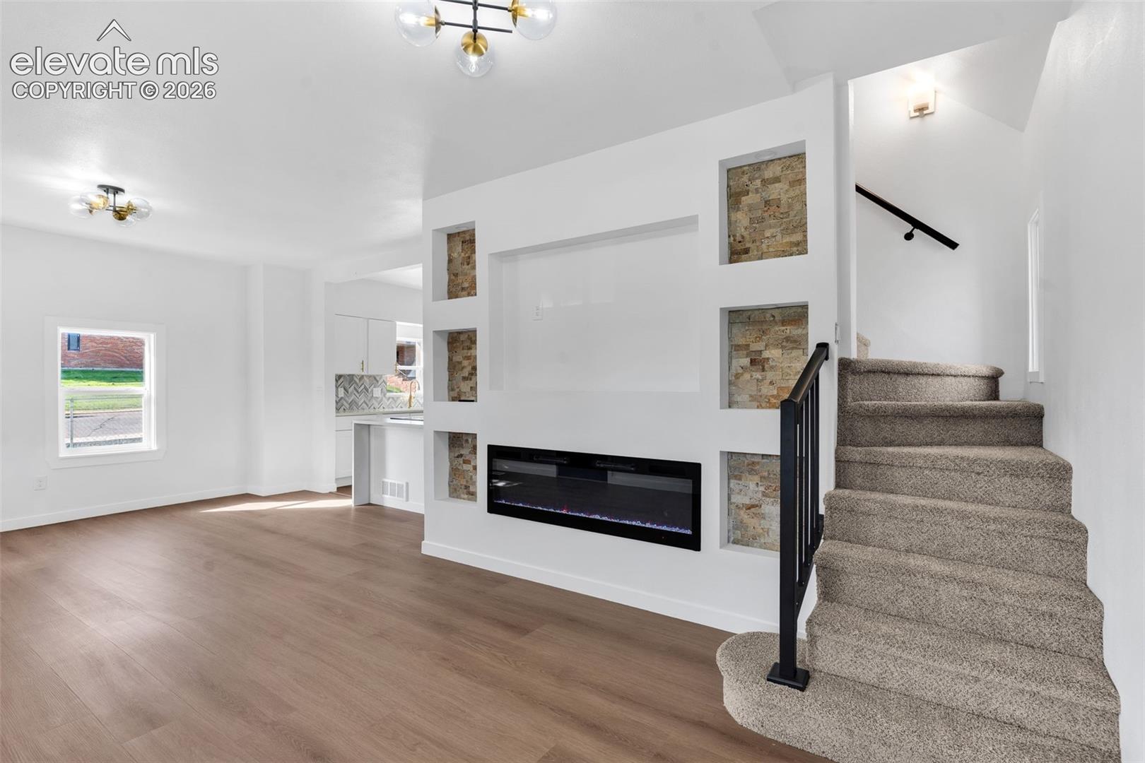 Living area featuring wood-finish flooring, a linear electric fireplace, stone-accented recessed shelving, and a carpeted staircase with a metal handrail