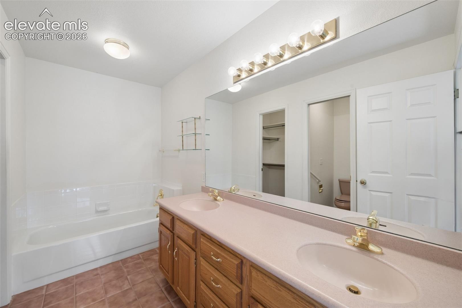 Dual-sink vanity with a solid-surface countertop and wood cabinetry, full-width wall mirror, multi-bulb vanity lighting, and a white soaking tub with integrated tile surround